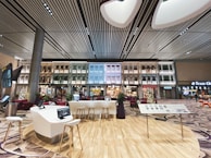 An indoor setting with a modern aesthetic, featuring a high, striped ceiling and walls that are designed to resemble traditional exterior building facades. The space includes contemporary seating arrangements, interactive screens, and digital kiosks. Christmas decorations, including a tree, are visible, suggesting a festive atmosphere.