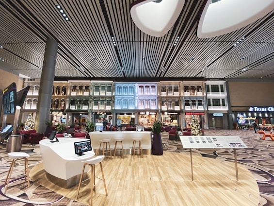 An indoor setting with a modern aesthetic, featuring a high, striped ceiling and walls that are designed to resemble traditional exterior building facades. The space includes contemporary seating arrangements, interactive screens, and digital kiosks. Christmas decorations, including a tree, are visible, suggesting a festive atmosphere.