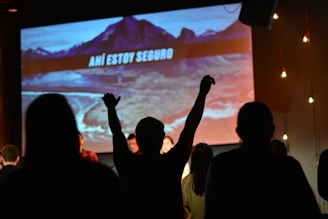 A group of people stands in a dimly lit room with a large screen displaying a scenic mountain image and the text 'AHÍ ESTOY SEGURO'. One person raises their arm in the air, possibly in excitement or worship.