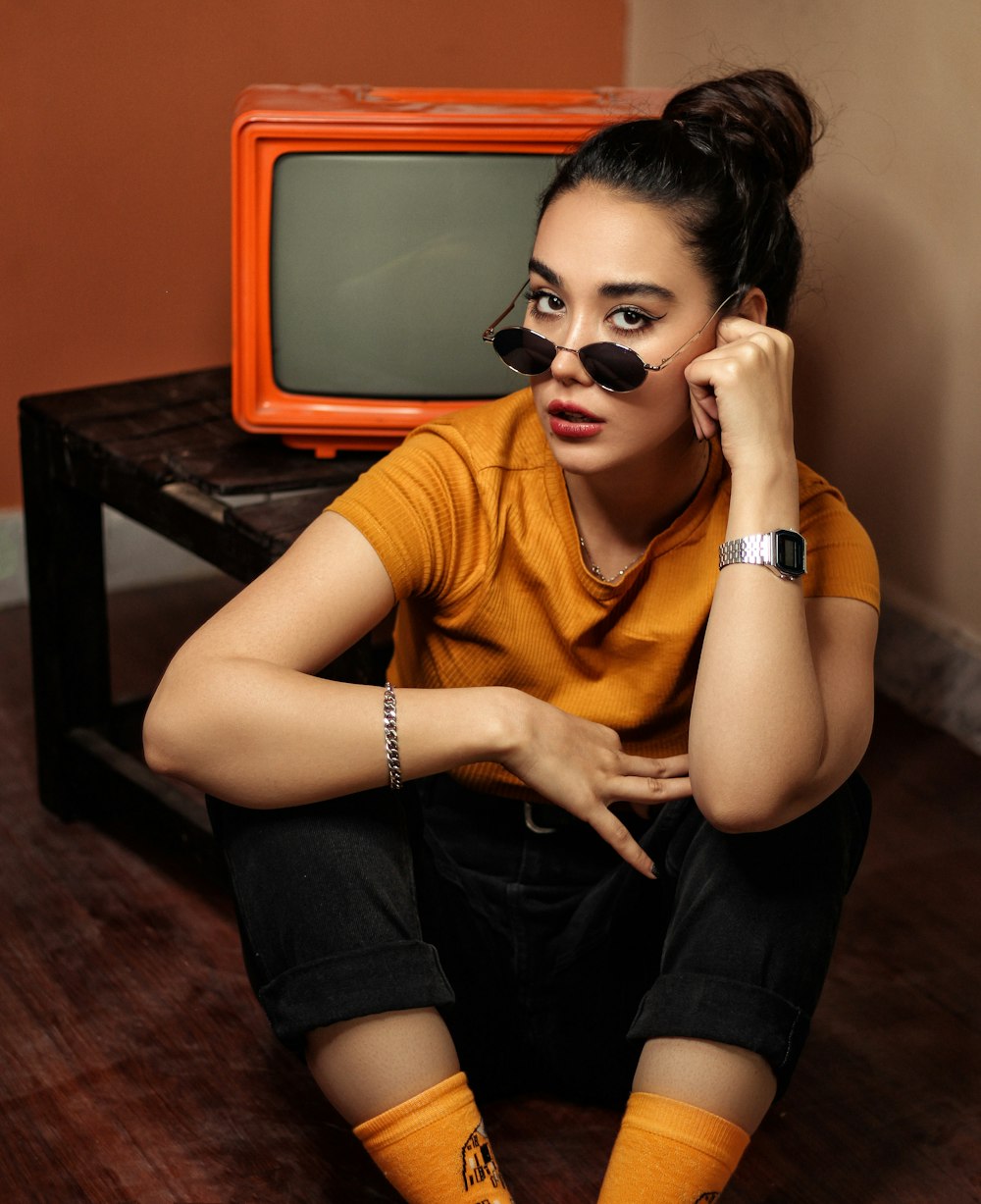 woman wearing orange crew-neck t-shirt, black denim jeans, and sunglasses sitting on floor near black wooden coffee table