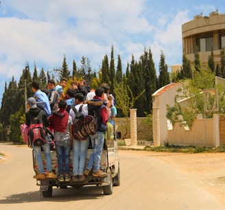 A group of children are standing on the back of a small truck, each carrying backpacks. The truck moves along a paved road lined with trees and residential buildings. The sky is partly cloudy, and tall evergreens are visible in the background.