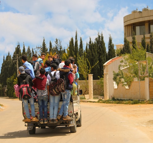 A group of children are standing on the back of a small truck, each carrying backpacks. The truck moves along a paved road lined with trees and residential buildings. The sky is partly cloudy, and tall evergreens are visible in the background.