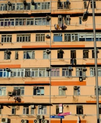 A multi-story residential building with numerous windows and balconies. Many air conditioning units are mounted on the walls, and clothes are hanging out to dry on some balconies. The building exterior is primarily orange and beige. A street lamp is visible, as well as street signs in the foreground.