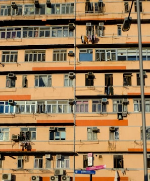 A multi-story residential building with numerous windows and balconies. Many air conditioning units are mounted on the walls, and clothes are hanging out to dry on some balconies. The building exterior is primarily orange and beige. A street lamp is visible, as well as street signs in the foreground.