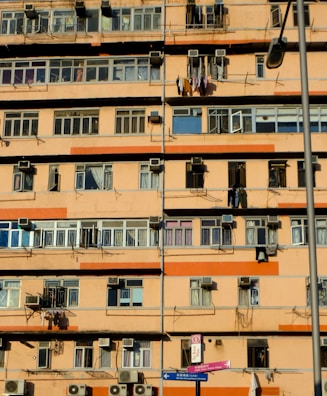 A multi-story residential building with numerous windows and balconies. Many air conditioning units are mounted on the walls, and clothes are hanging out to dry on some balconies. The building exterior is primarily orange and beige. A street lamp is visible, as well as street signs in the foreground.