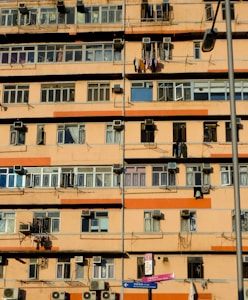 A multi-story residential building with numerous windows and balconies. Many air conditioning units are mounted on the walls, and clothes are hanging out to dry on some balconies. The building exterior is primarily orange and beige. A street lamp is visible, as well as street signs in the foreground.