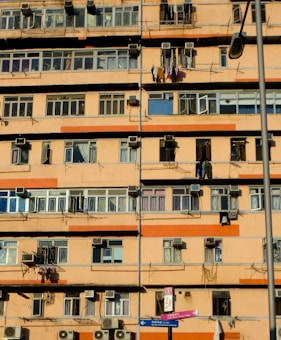 A multi-story residential building with numerous windows and balconies. Many air conditioning units are mounted on the walls, and clothes are hanging out to dry on some balconies. The building exterior is primarily orange and beige. A street lamp is visible, as well as street signs in the foreground.