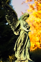 A stone statue of an angel with large, intricate wings stands gracefully in a natural setting. The angel's gaze is downward and serene, with its hands gently clasped. The background features vibrant autumn foliage with warm orange and yellow hues, contrasting against the dark green of surrounding bushes.