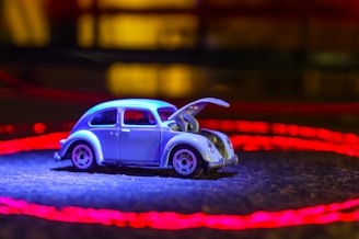 A vintage toy car, reminiscent of a classic Volkswagen Beetle, is placed on a surface with its hood open. The car is illuminated by a bluish light, while a vibrant red circle of light surrounds it. The background is dimly lit, creating a contrast with the brightly lit toy car.