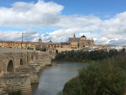 A historical stone bridge arches over a river, leading to a grand architectural structure in the background. The skyline includes multifaceted domes and towers rising above other buildings. A tall crane can be seen on the left side, indicating construction activity. The foreground features lush greenery alongside the riverbank, with mountains visible far away under a partly cloudy sky.