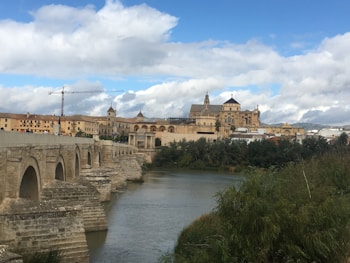 A historical stone bridge arches over a river, leading to a grand architectural structure in the background. The skyline includes multifaceted domes and towers rising above other buildings. A tall crane can be seen on the left side, indicating construction activity. The foreground features lush greenery alongside the riverbank, with mountains visible far away under a partly cloudy sky.
