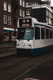 A tram with the route number 19 heading to Station Sloterdijk travels along a street. The tram is white with blue accents and displays bright front lights. Surrounding the tram are several multi-story buildings with numerous windows. A shop with a bright sign is visible on the left, and the infrastructure features tram tracks integrated into the road.