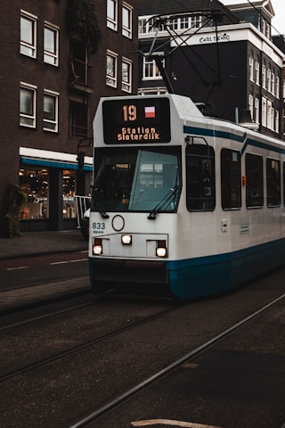 A tram with the route number 19 heading to Station Sloterdijk travels along a street. The tram is white with blue accents and displays bright front lights. Surrounding the tram are several multi-story buildings with numerous windows. A shop with a bright sign is visible on the left, and the infrastructure features tram tracks integrated into the road.