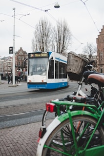 A light rail tram is on the street, showing the destination 'Nieuw Sloten'. In the foreground, a bicycle with a basket is parked, and tram tracks are visible on the road. Buildings and leafless trees are present in the background, with overhead tram wires crossing the sky.