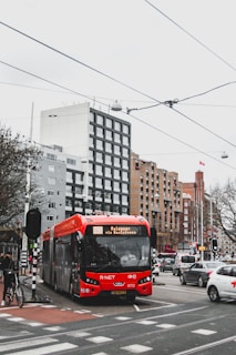 A red public bus is on a city street, surrounded by cars and bicycles. Several modern buildings with various architectural designs are visible in the background. Overhead wires and traffic lights are present, and a few pedestrians are walking nearby.