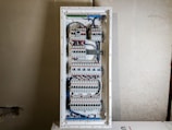 A large electrical panel with multiple circuit breakers, wires, and switches neatly arranged inside a rectangular metal casing. The wires are mainly blue and white, connecting different components. The panel is mounted on a plain, unfinished wall.