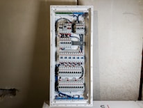 A large electrical panel with multiple circuit breakers, wires, and switches neatly arranged inside a rectangular metal casing. The wires are mainly blue and white, connecting different components. The panel is mounted on a plain, unfinished wall.