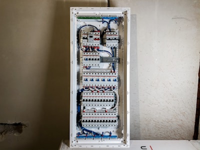 A large electrical panel with multiple circuit breakers, wires, and switches neatly arranged inside a rectangular metal casing. The wires are mainly blue and white, connecting different components. The panel is mounted on a plain, unfinished wall.
