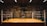 An empty boxing ring is set up in a dimly lit gym with various boxing posters adorning the walls. The ropes of the ring are slightly worn, and the space has an aged, rustic feel.