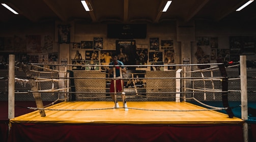 An empty boxing ring is set up in a dimly lit gym with various boxing posters adorning the walls. The ropes of the ring are slightly worn, and the space has an aged, rustic feel.