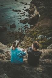 two women sitting on cliff while chatting viewing blue body of water