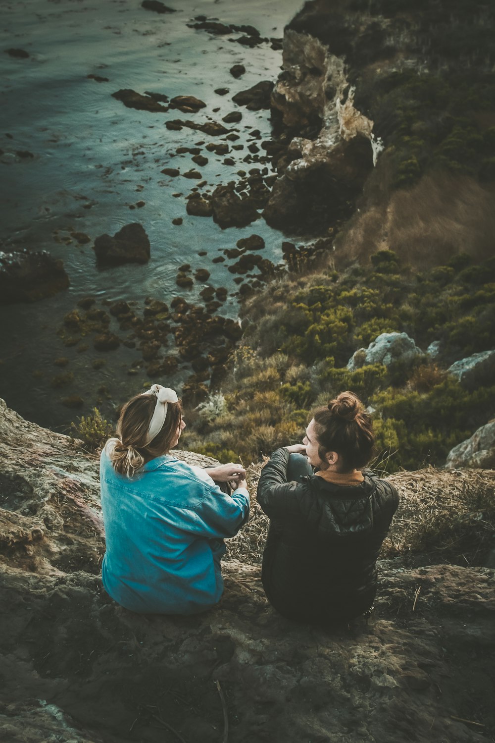 two women sitting on cliff while chatting viewing blue body of water