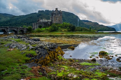 A historic castle sits majestically on the edge of a reflective loch, surrounded by rugged rocks and lush greenery. The sky is overcast with thick clouds, adding a dramatic aura to the scene. In the foreground, patches of vibrant moss and grass are interspersed among the rocky terrain.