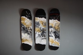 Three skateboard decks are mounted on a wall, featuring abstract art with swirling patterns in black, white, and gold. The designs include bold, sweeping brushstrokes and intricate splatters that create a dynamic, fluid appearance.
