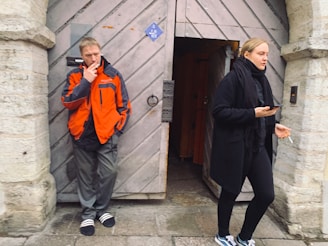A man in an orange and black jacket is leaning against a stone wall, smoking a cigarette. He stands to the left of a wooden door that is slightly ajar. A woman in a black coat and scarf is walking past on the right, holding a phone in one hand and a cigarette in the other.