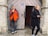A man in an orange and black jacket is leaning against a stone wall, smoking a cigarette. He stands to the left of a wooden door that is slightly ajar. A woman in a black coat and scarf is walking past on the right, holding a phone in one hand and a cigarette in the other.