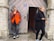 A man in an orange and black jacket is leaning against a stone wall, smoking a cigarette. He stands to the left of a wooden door that is slightly ajar. A woman in a black coat and scarf is walking past on the right, holding a phone in one hand and a cigarette in the other.