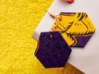 Two hexagonal-shaped fabric earrings with bold patterns, one predominantly purple with black designs and the other bright yellow with detailed black designs, are placed on a textured yellow-gold surface.