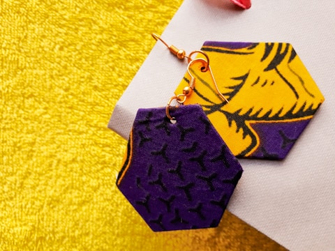 Two hexagonal-shaped fabric earrings with bold patterns, one predominantly purple with black designs and the other bright yellow with detailed black designs, are placed on a textured yellow-gold surface.