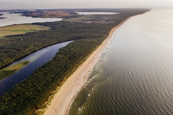 Aerial view showcasing an expansive coastline with a long sandy beach on the right, bordered by the sea with gentle waves and a forested area on the left. Inland, a mix of forests, rivers, and small towns are visible, along with distant fields and other water bodies.