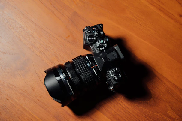 A close-up of a sleek digital camera resting on a wooden table with natural light highlighting its lens.
