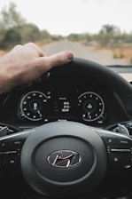 A hand is placed on the steering wheel of a Hyundai car, with a view of the dashboard displaying digital gauges and controls. The road ahead is visible through the windshield, surrounded by blurred trees.