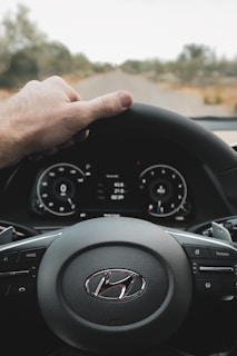 A hand is placed on the steering wheel of a Hyundai car, with a view of the dashboard displaying digital gauges and controls. The road ahead is visible through the windshield, surrounded by blurred trees.
