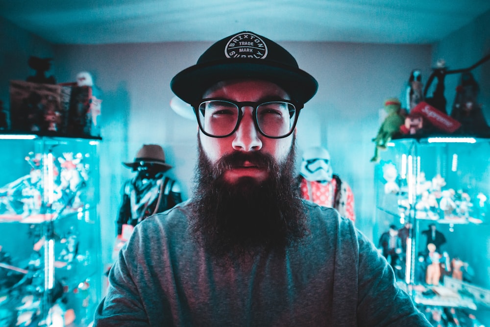 man wearing gray long-sleeved shirt, black and white hat, and eyeglasses inside green room