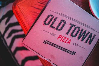 A pizza box labeled 'Old Town Pizza' placed on a wooden surface with a patterned cloth partially visible beneath. The text on the box indicates 'Handcrafted Since 1998' in a stylized font.