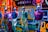 Mannequins displaying casual clothing are positioned inside a vibrant store window. The background is filled with colorful neon signs and lights, creating a bright and dynamic atmosphere. The mannequins are dressed in shorts and polo shirts, showcasing different styles and colors.
