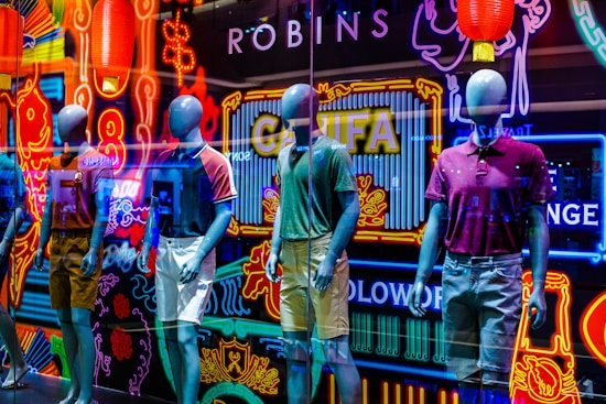 Mannequins displaying casual clothing are positioned inside a vibrant store window. The background is filled with colorful neon signs and lights, creating a bright and dynamic atmosphere. The mannequins are dressed in shorts and polo shirts, showcasing different styles and colors.