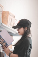 A person wearing a black cap and black shirt reads a book while standing next to a shelf filled with various books. The setting appears to be a library or bookstore with a relaxed and quiet ambiance.