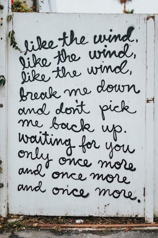 A white wooden fence with black cursive writing displaying a poetic message. The text reads: 'like the wind, like the wind, like the wind, break me down and don't pick me back up. waiting for you only once more and once more and once more.' There are small plants growing around the fence.