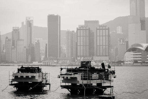 Two large floating platforms sit on the water, tethered by ropes. They are positioned in front of a cityscape dominated by tall skyscrapers and modern buildings. The ambiance is misty or hazy, which softens the outlines of the distant structures.