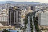 A bustling urban landscape featuring a high-rise building under construction in the foreground, surrounded by numerous apartment complexes and commercial buildings. A busy street with cars runs through the city, lined with trees on both sides. The skyline is dotted with cranes, indicating ongoing development, against a backdrop of distant mountains.