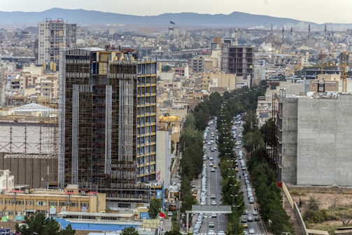 A bustling urban landscape featuring a high-rise building under construction in the foreground, surrounded by numerous apartment complexes and commercial buildings. A busy street with cars runs through the city, lined with trees on both sides. The skyline is dotted with cranes, indicating ongoing development, against a backdrop of distant mountains.