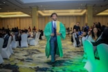 A person in a green and yellow graduation gown walks confidently down the aisle of a well-lit conference room. The room is filled with people seated around tables, dressed in formal attire. The carpet features a multicolored pattern, and the ceiling is elegantly designed with chandeliers. Several attendees are watching or capturing the moment with cameras.