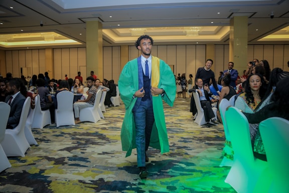 A person in a green and yellow graduation gown walks confidently down the aisle of a well-lit conference room. The room is filled with people seated around tables, dressed in formal attire. The carpet features a multicolored pattern, and the ceiling is elegantly designed with chandeliers. Several attendees are watching or capturing the moment with cameras.