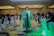 A person in a green and yellow graduation gown walks confidently down the aisle of a well-lit conference room. The room is filled with people seated around tables, dressed in formal attire. The carpet features a multicolored pattern, and the ceiling is elegantly designed with chandeliers. Several attendees are watching or capturing the moment with cameras.