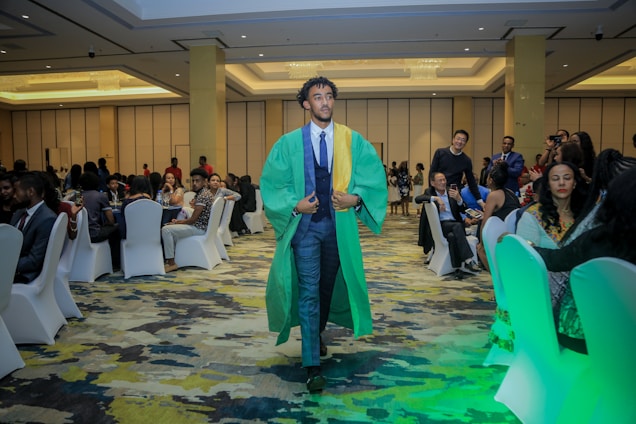 A person in a green and yellow graduation gown walks confidently down the aisle of a well-lit conference room. The room is filled with people seated around tables, dressed in formal attire. The carpet features a multicolored pattern, and the ceiling is elegantly designed with chandeliers. Several attendees are watching or capturing the moment with cameras.